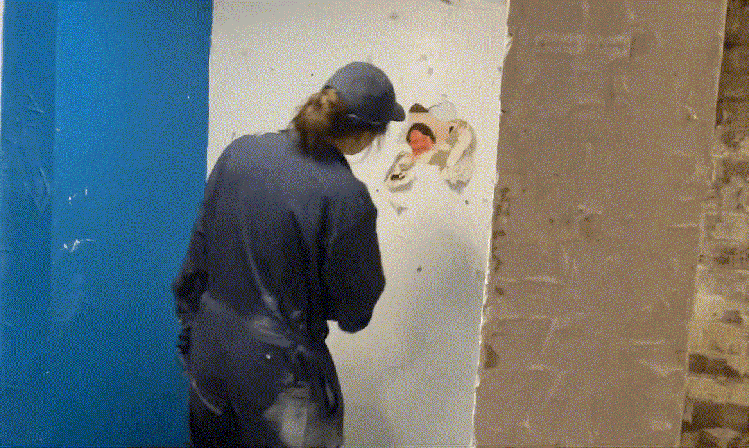A woman waves through a hole in the wall and then shakes hands with someone on the other side through it.