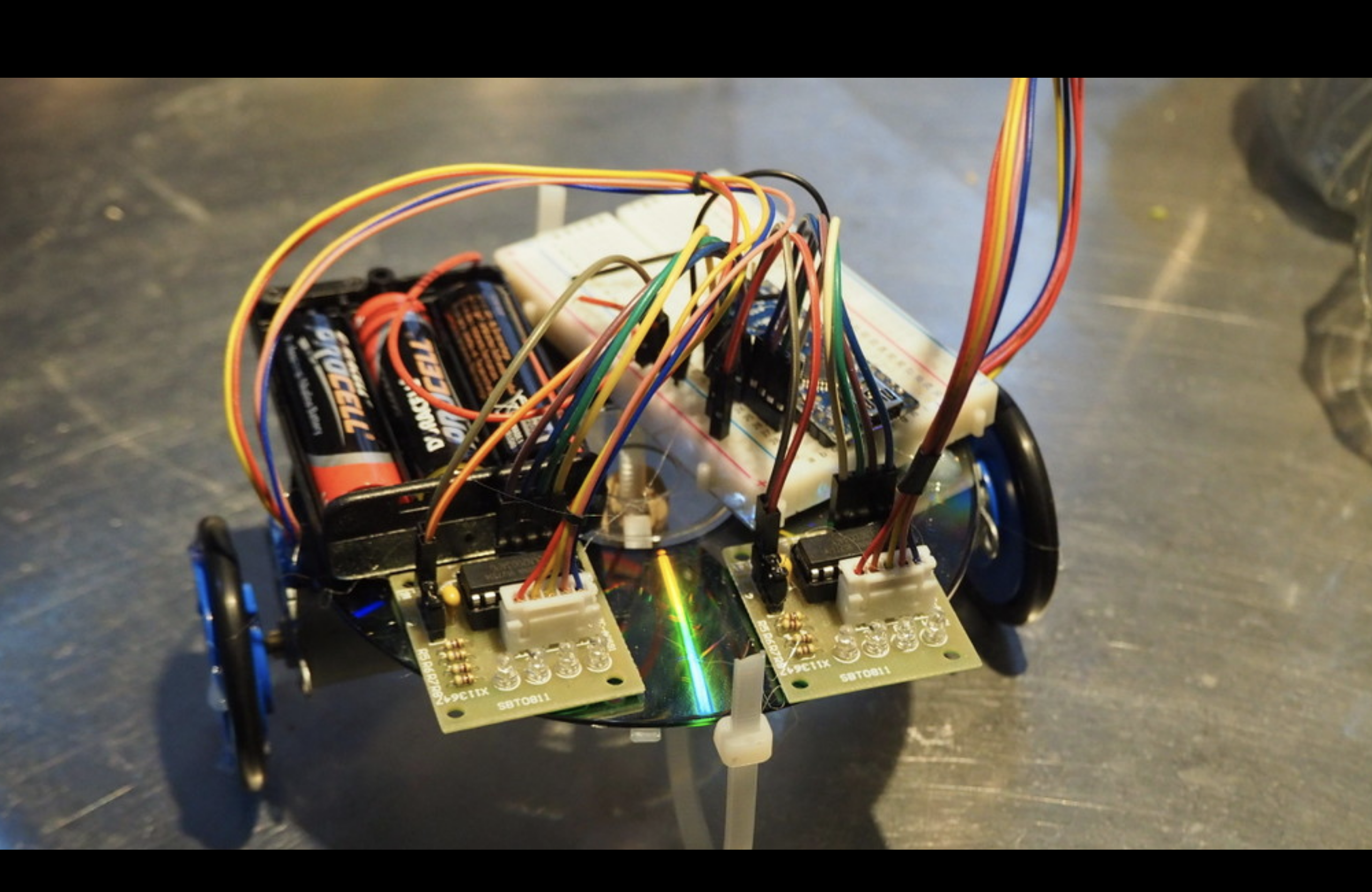 A close up of shonkbot on a shiny table. You can see it's made of a CD, plus some motors, an arduino nano, a breadboard and an AA battery back.