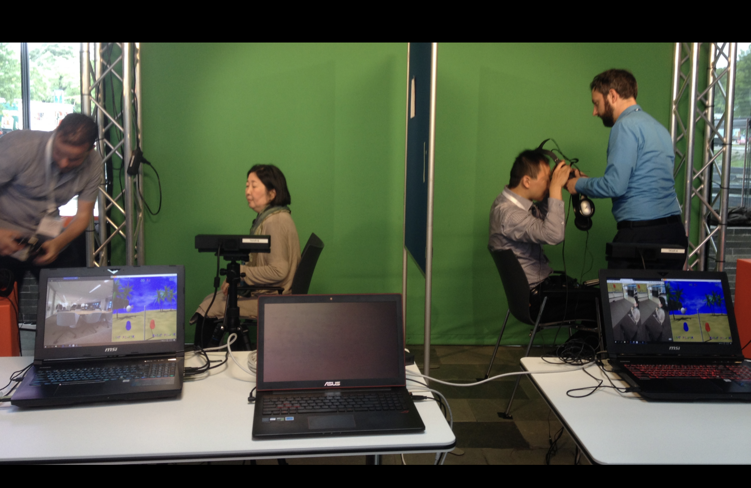 Two people getting helped to wear VR headsets against a green background